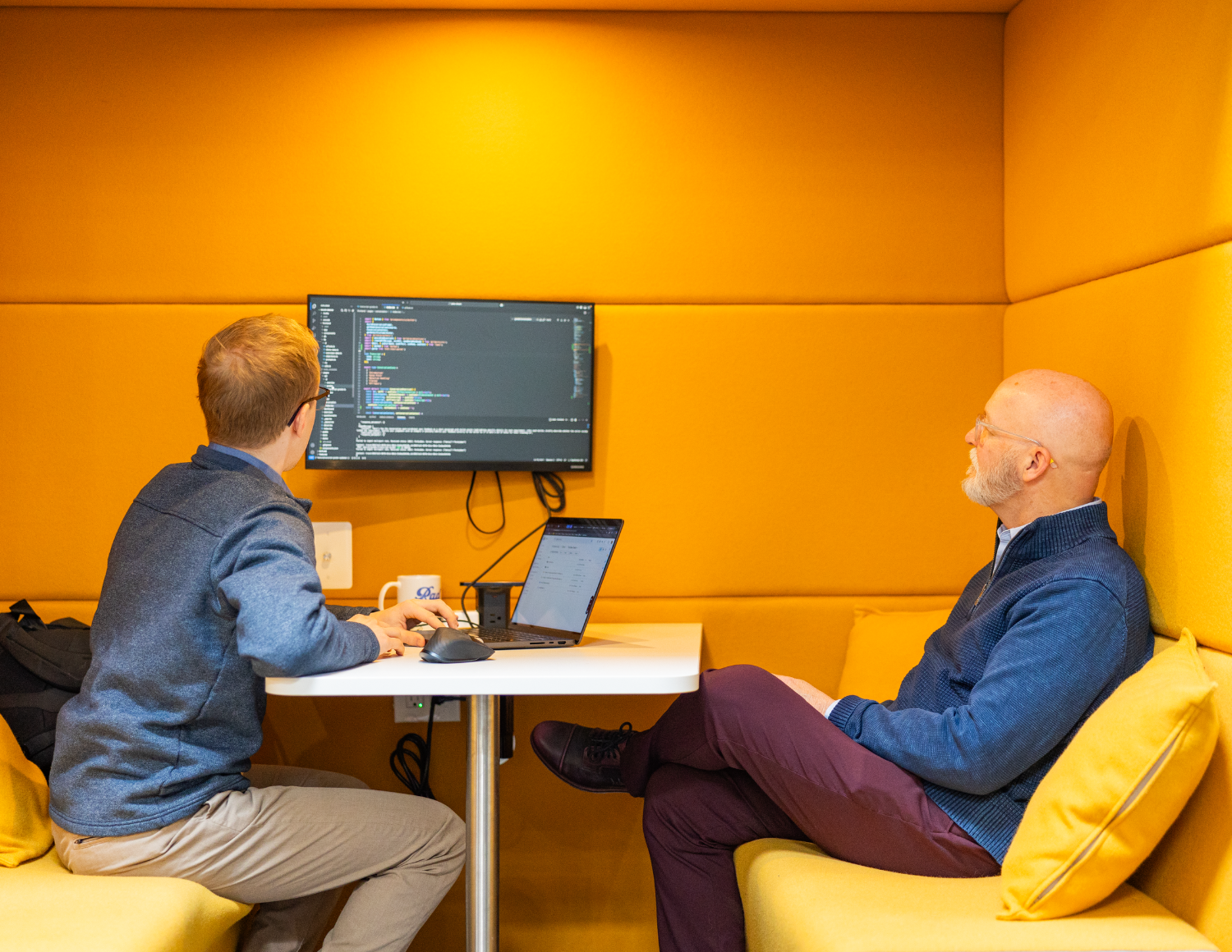 Two people sit in a cozy meeting booth with yellow cushioned walls. One points at a screen displaying code, while the other listens intently. A laptop and coffee cups are on the table, hinting at a tech discussion or collaboration for their data solutions company.