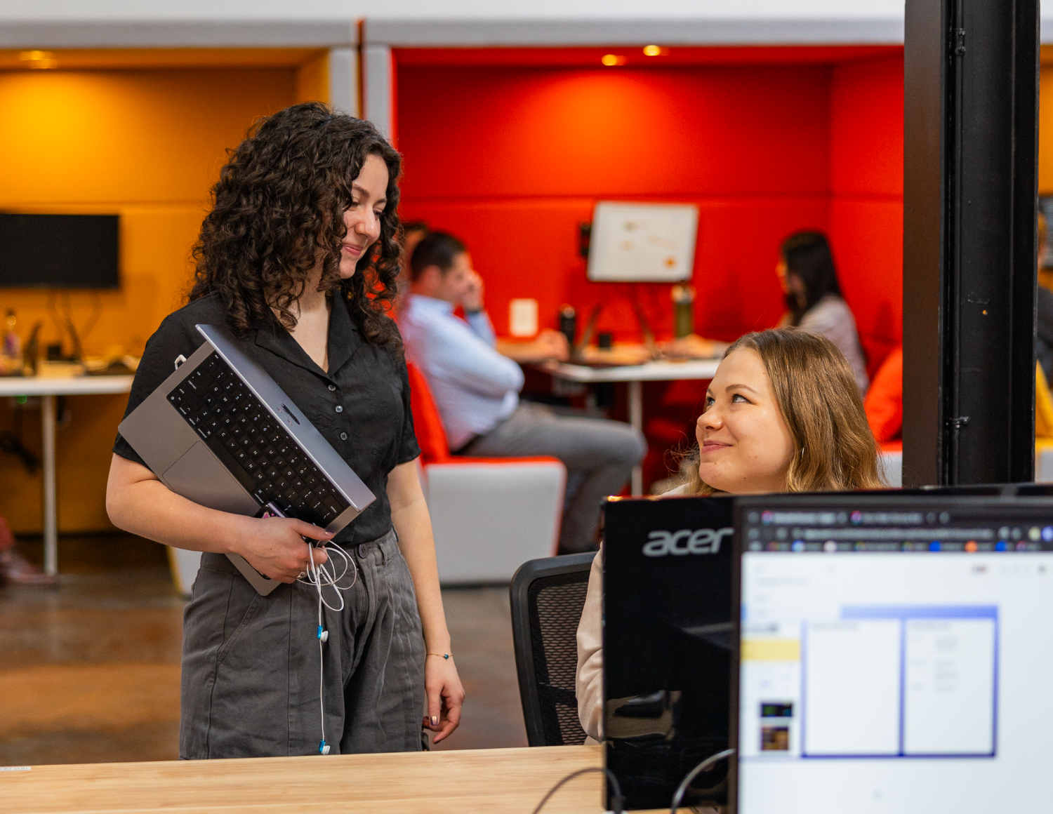 Two women in an office setting at a data solutions company—one standing with a laptop, smiling at the seated woman. Others diligently work in the background at vibrant orange and red workstations.