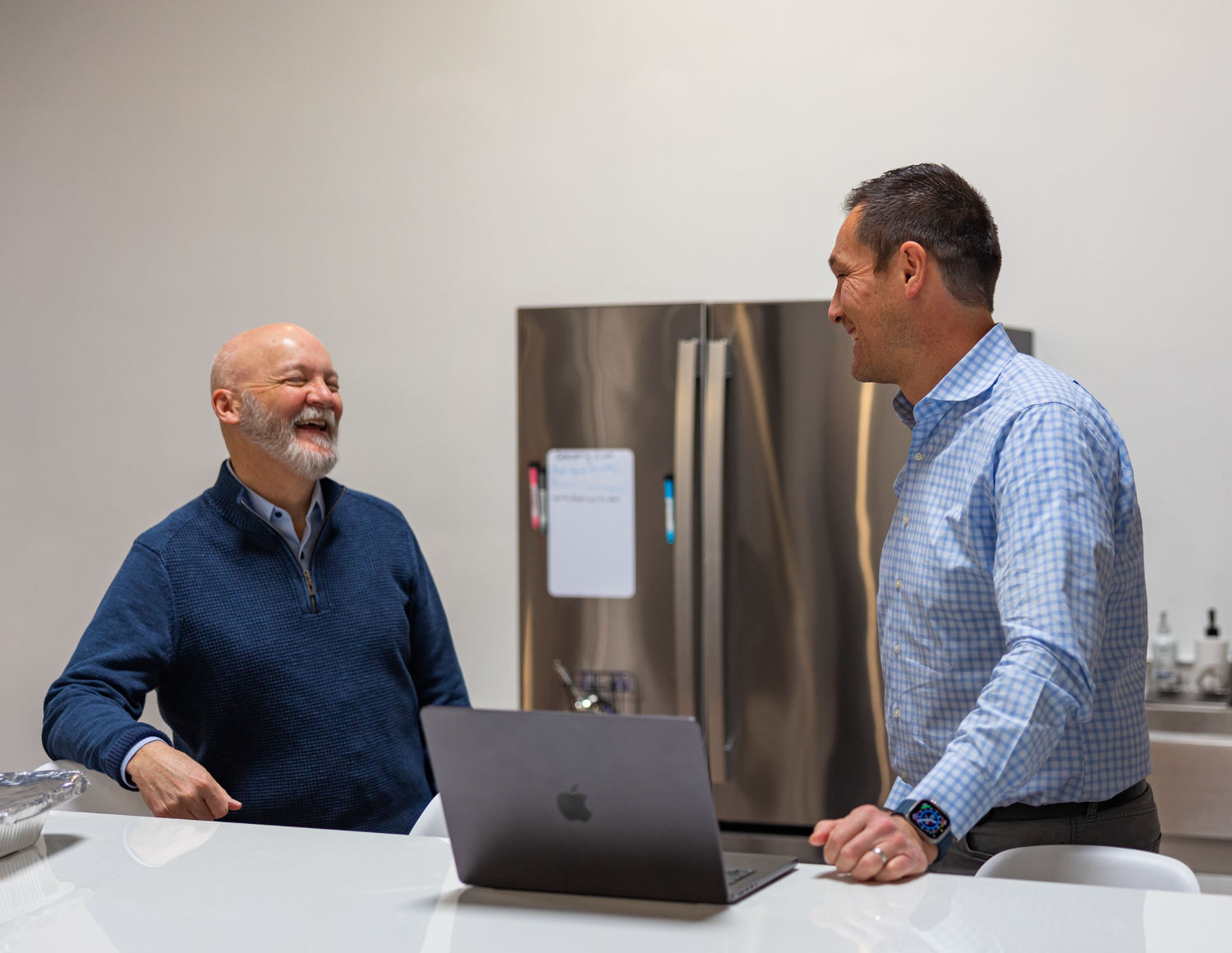 Two men are standing in a kitchen area, smiling and discussing AI strategy. The bald man with a beard is in a dark sweater, while the other, sporting short hair, wears a light plaid shirt. They stand around an open laptop on the white countertop, with a fridge in the background.