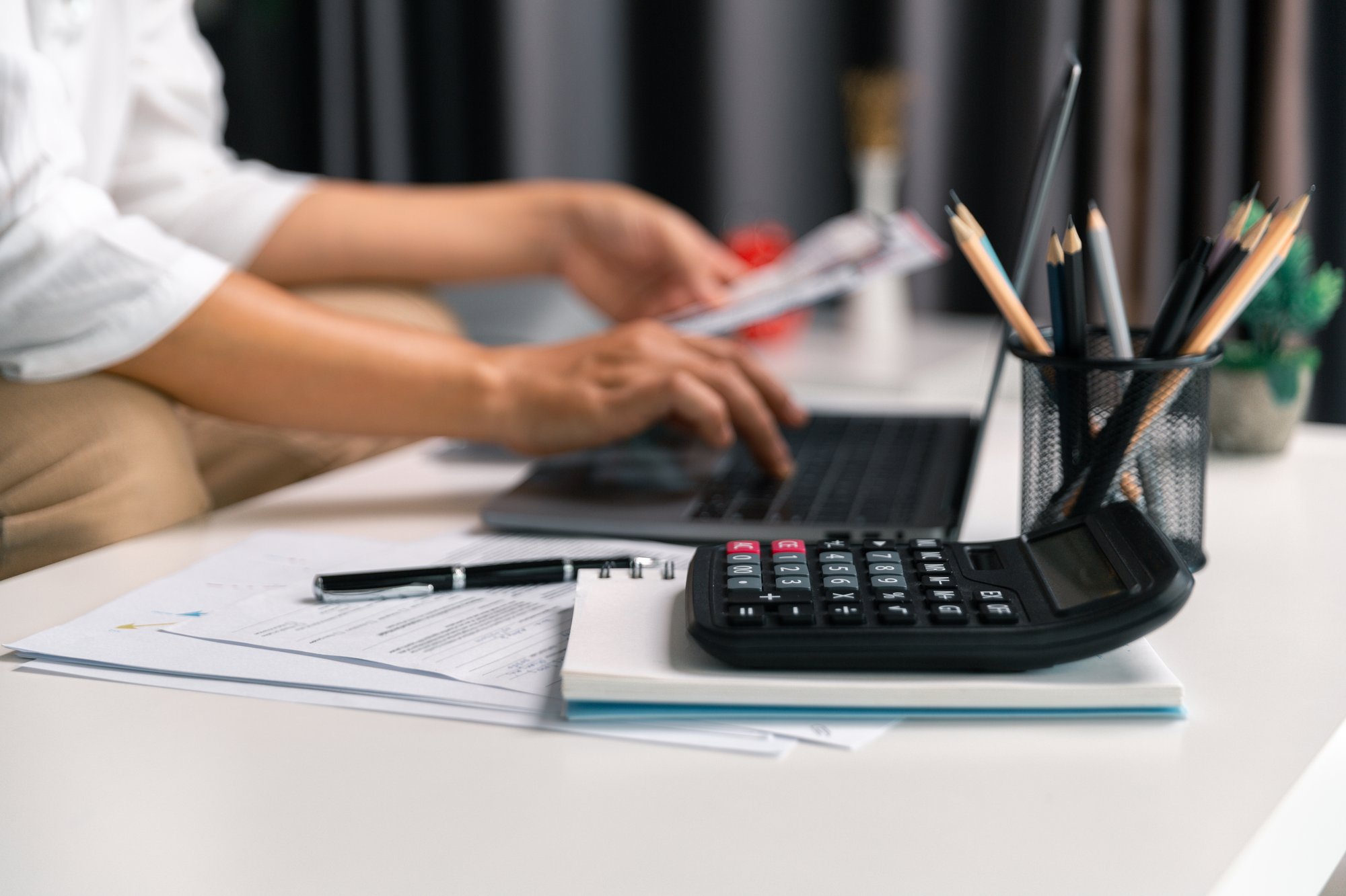 A person uses a laptop at a desk with documents, a pen, a calculator, and a container holding pencils and pens, suggesting work on finances or office tasks—possibly reviewing private equity tech solutions.