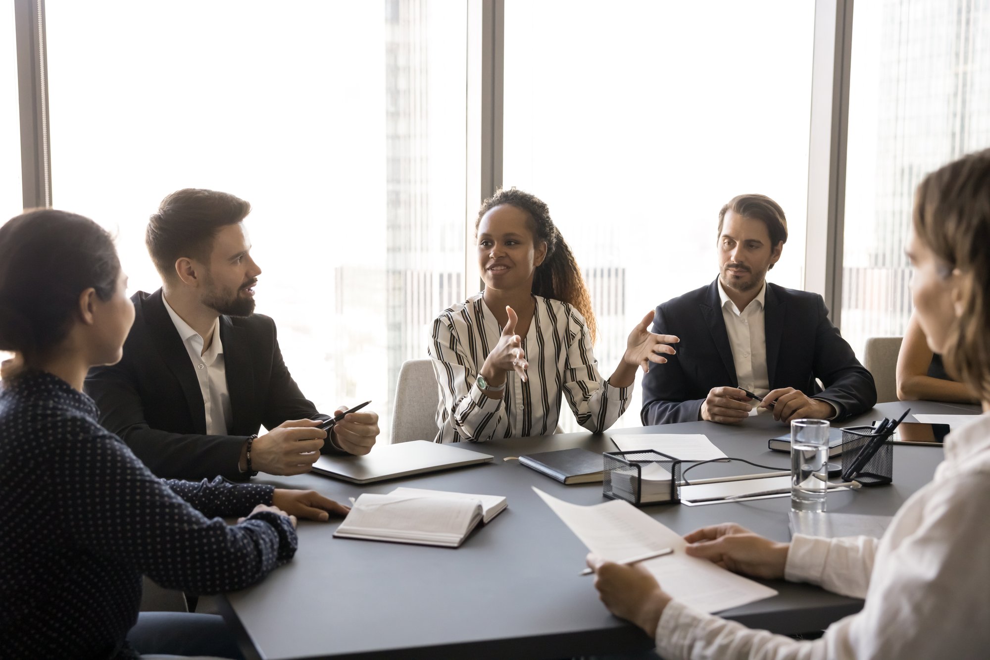 Five people sit around a conference table in an office, engaged in discussion about AI for private equity. One woman gestures as she speaks while others listen and take notes. Laptops, papers, and a glass of water are on the table.