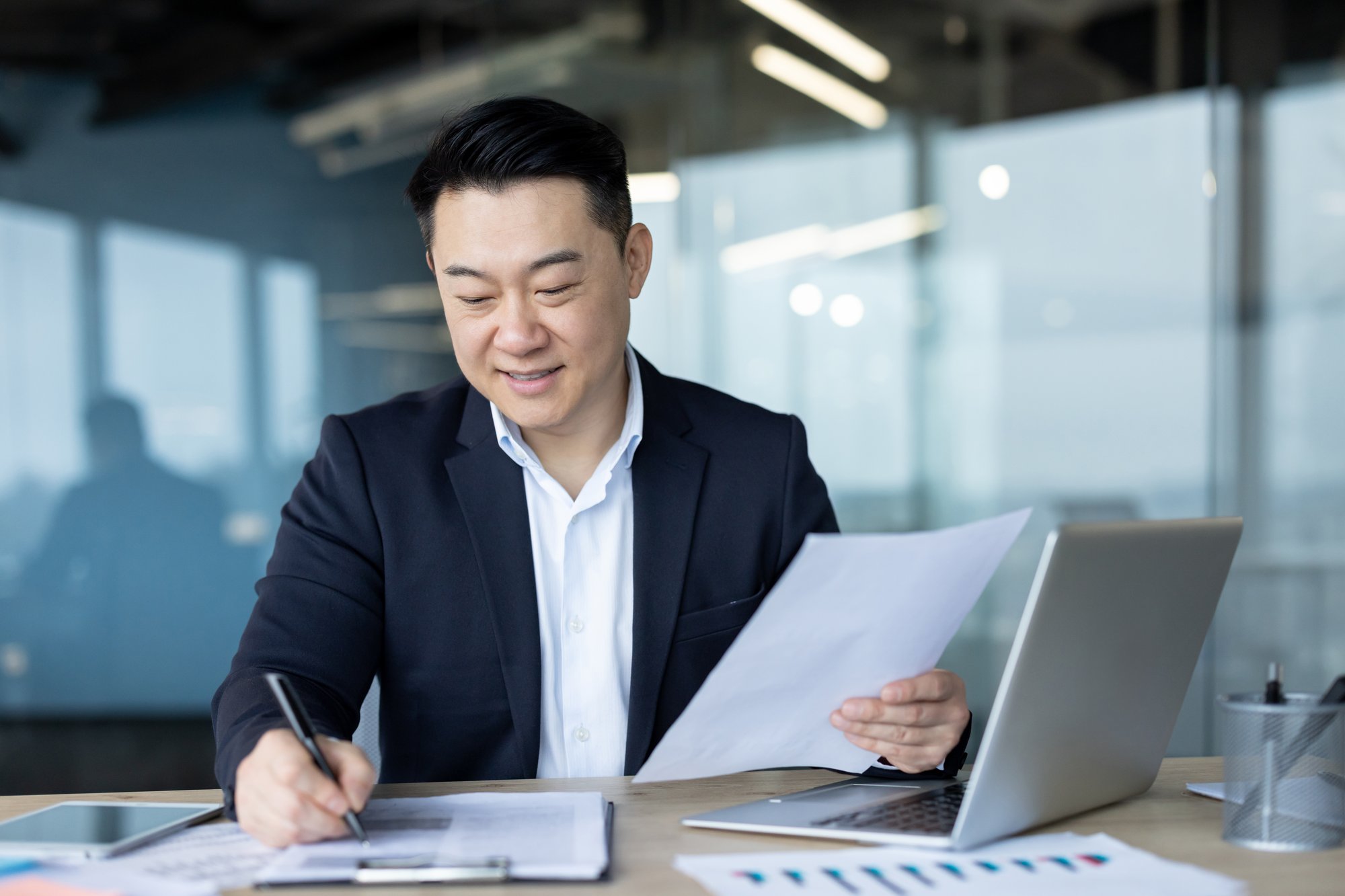 A man in a suit sits at a desk with a laptop, holding papers and smiling while writing notes. Office environment with glass walls and documents visible, highlighting the use of private equity tech solutions.
