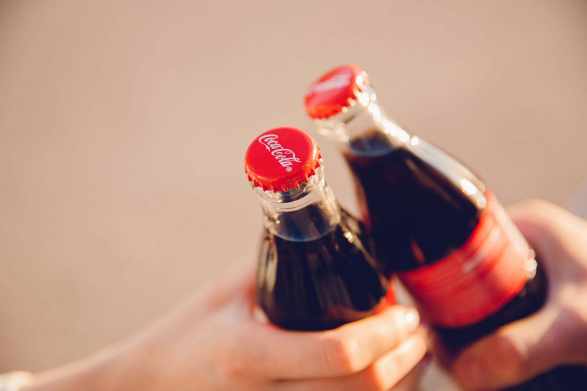 Two hands clink classic glass bottles of Coca-Cola, red caps gleaming. The softly blurred background highlights the focus on the user experience of this celebratory moment.