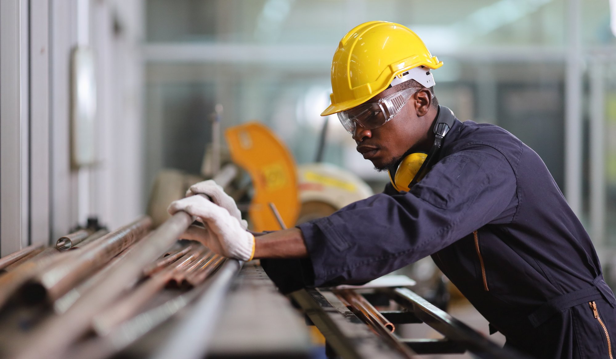 A male factory worker in a yellow hard hat, safety goggles, gloves, and coveralls inspects metal pipes on a workbench, demonstrating how AI for private equity can enhance efficiency in industrial settings.