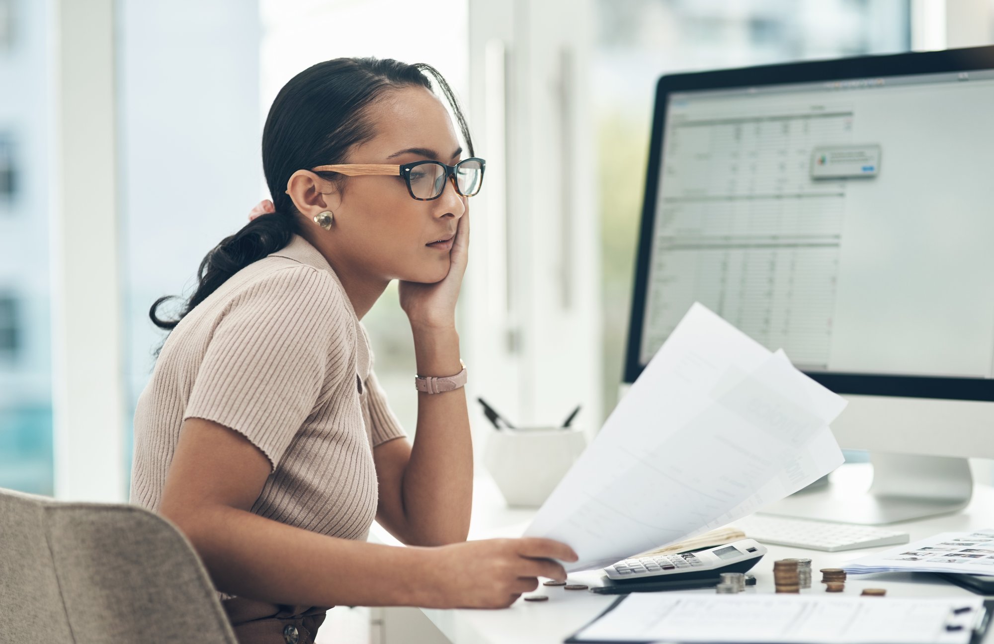 A woman wearing glasses sits at a desk, looking at papers in her hand. A calculator, coins, and documents are on the desk, with a large monitor displaying a spreadsheet featuring private equity tech solutions in the background.
