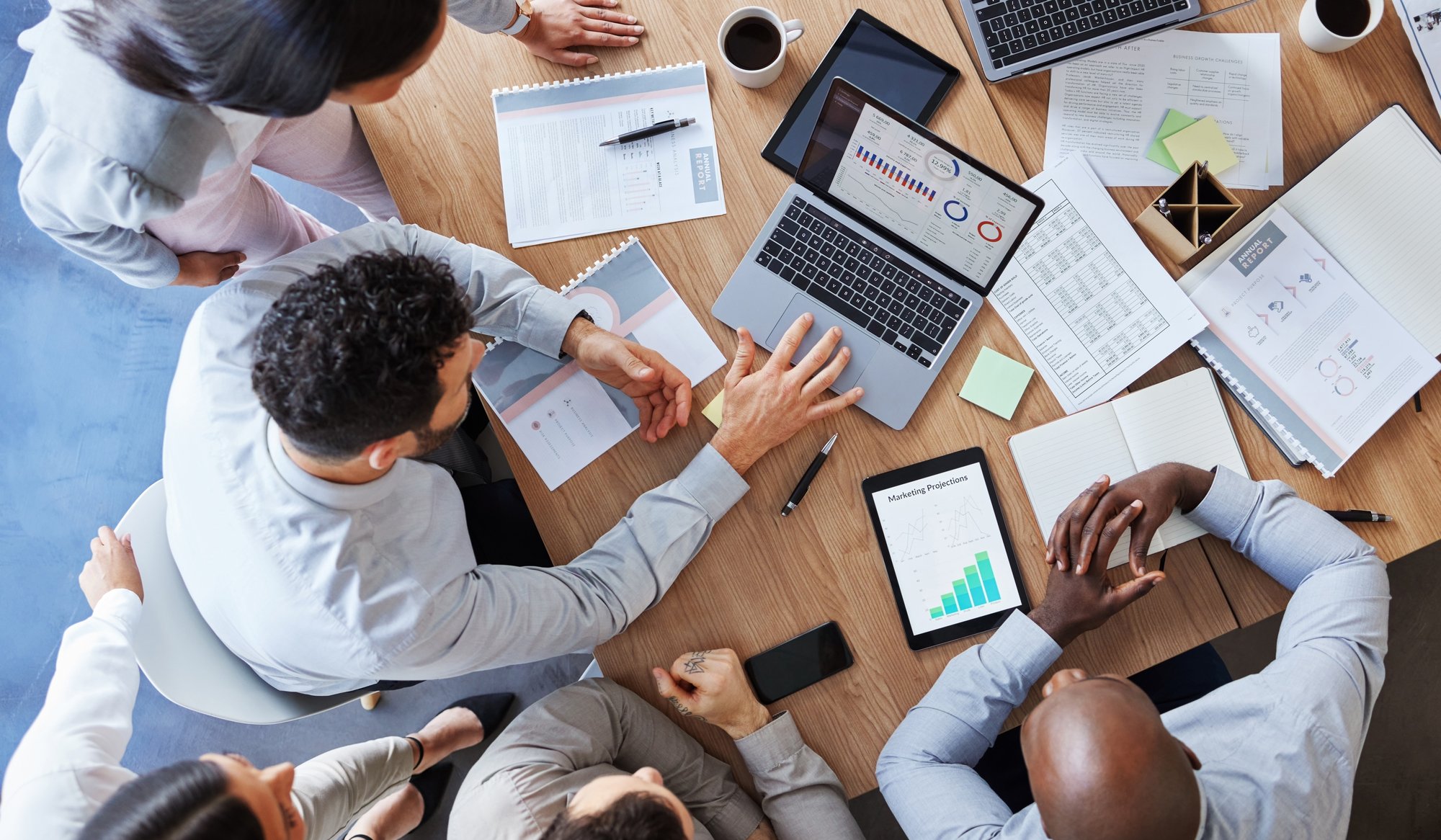 A group of people sit around a wooden table with laptops, tablets, and charts, collaborating on work. One person points at a laptop screen displaying graphs and data powered by AI for private equity.