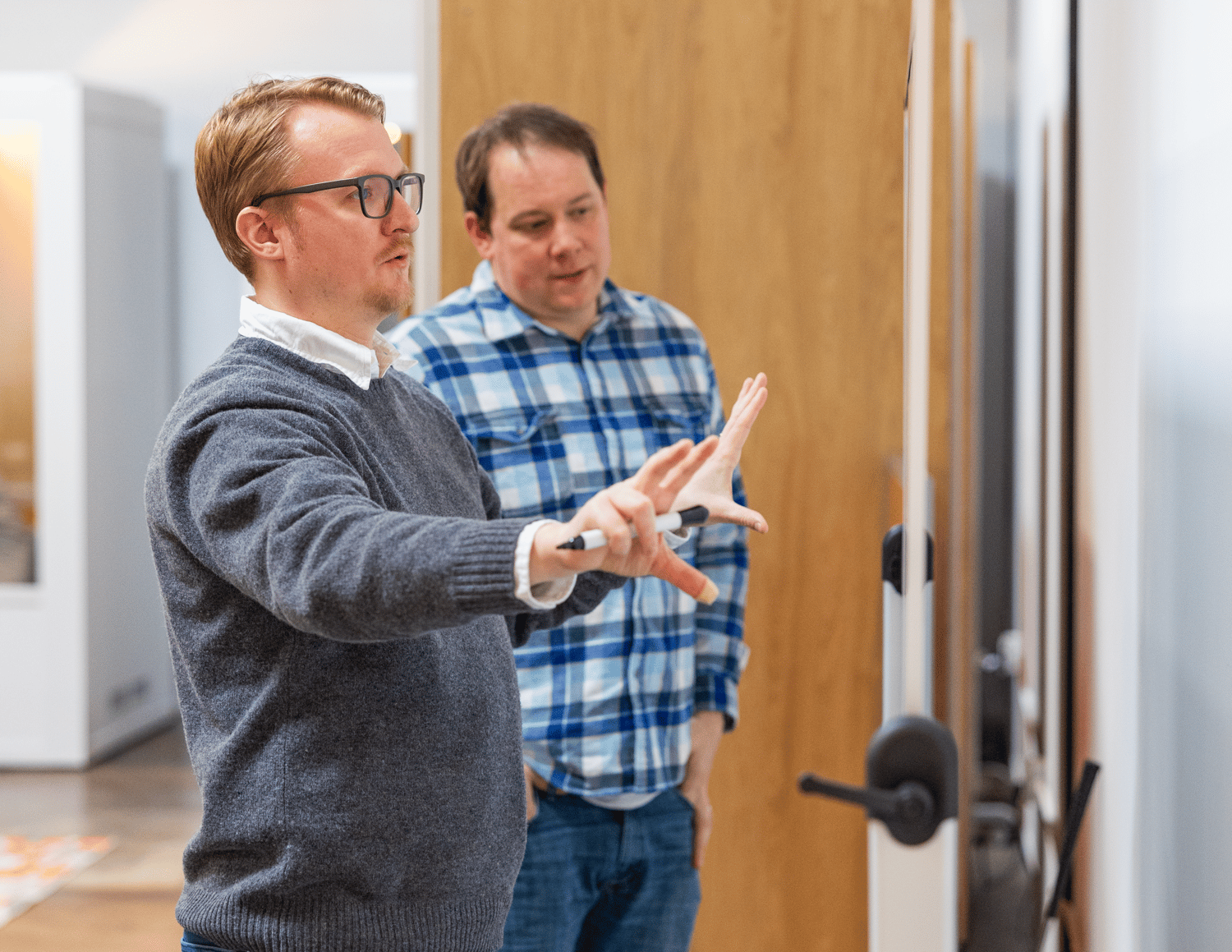 Two men in a modern office setting, engaged in a software consulting discussion. One wears a gray sweater and glasses, gesturing with his hands while holding a marker. The other wears a blue plaid shirt, attentively observing. A large whiteboard is in the background.