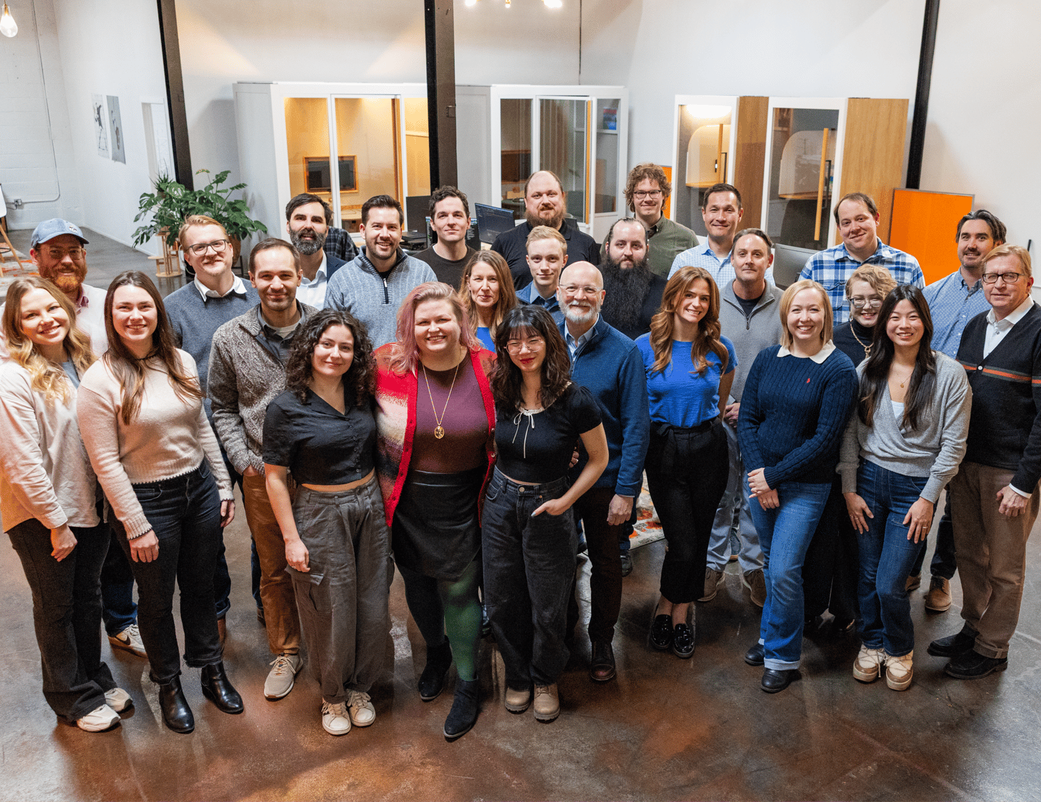 A diverse group of people stands together in a modern, open-plan office space with glass-walled meeting rooms in the background. The team, representing leading MN software companies, consists of smiling men and women dressed casually, showcasing a collaborative work environment.