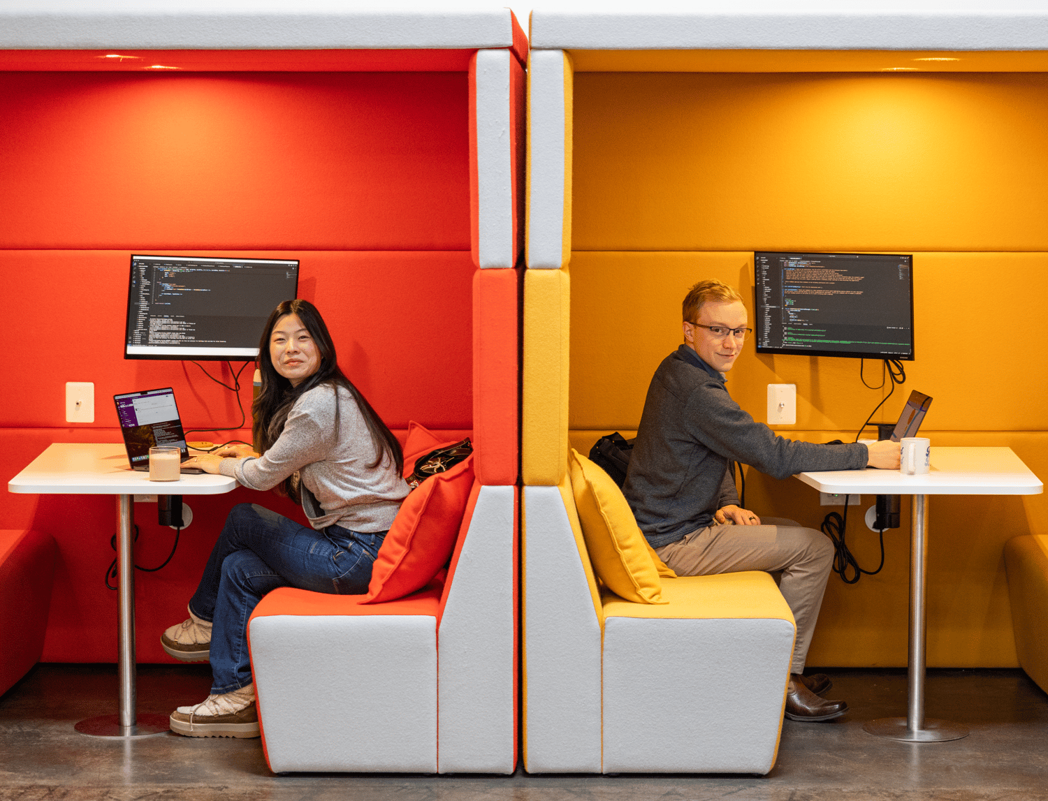 Two people sit back-to-back in vibrant booths, immersed in software consulting. The left booth is red, with a woman diligently typing on her laptop. The right booth is yellow, where a man intently focuses on his screen. Both have intricate code displayed on their monitors.