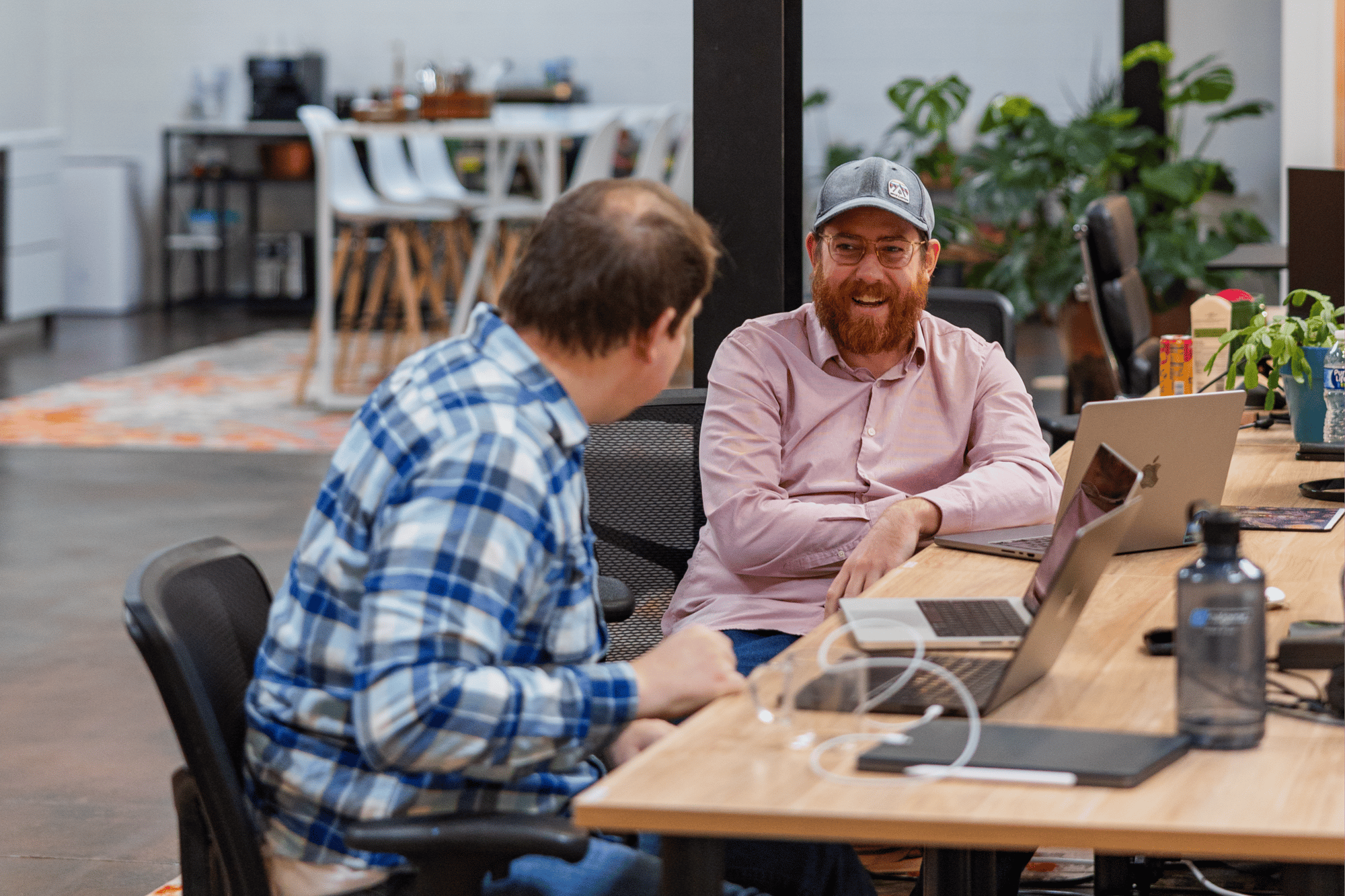 Two men sit at a shared desk in a modern office, talking and smiling. One wears a blue plaid shirt, the other a pink shirt and cap. Laptops, water bottles, and plants fill the background as they discuss AI for private equity solutions.