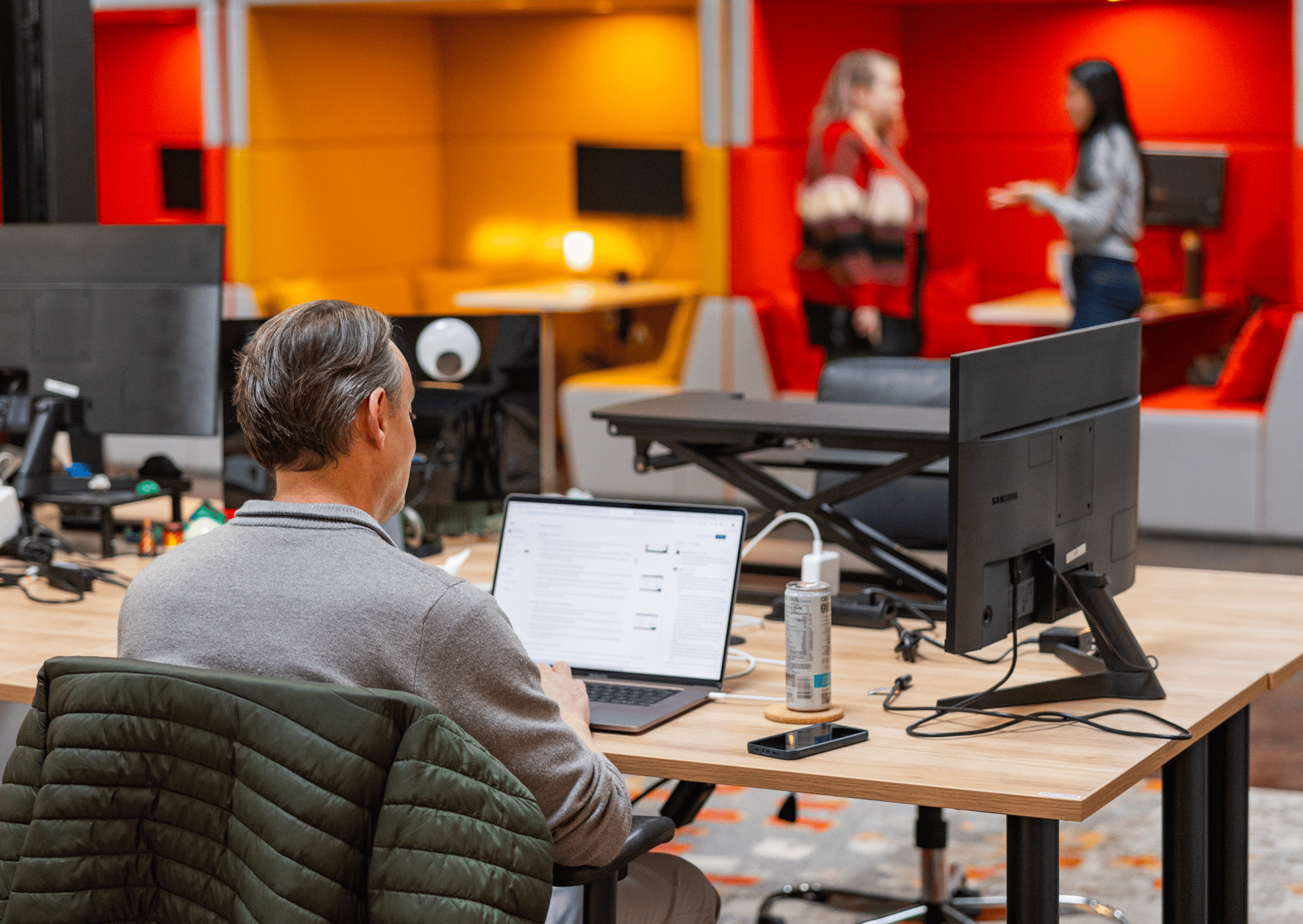 A man works at a desk on a laptop in a modern office, while two women talk in the colorful, blurred background. Desks and monitors hint at AI for private equity and tech solutions throughout the vibrant workspace.