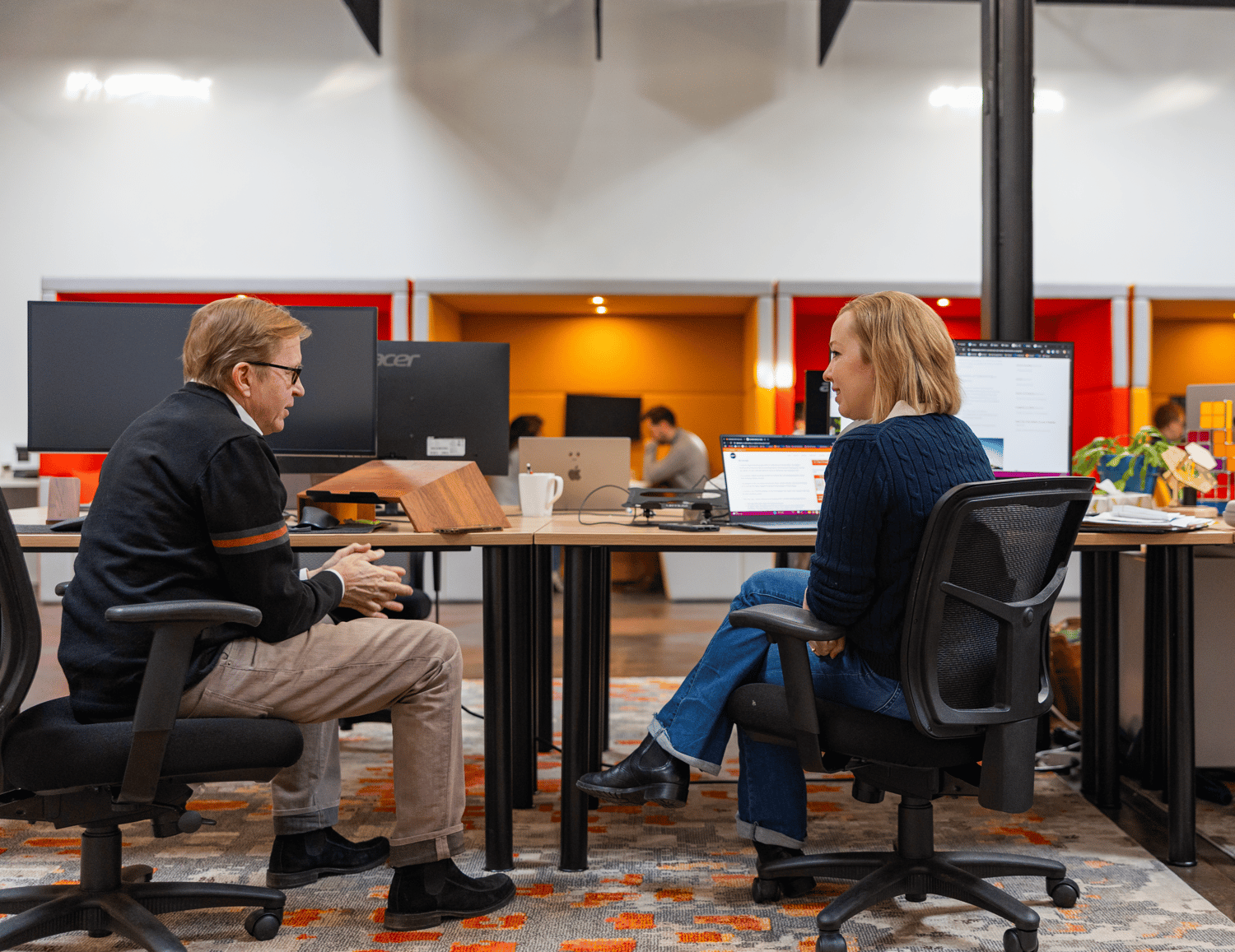 Two people sitting at desks with laptops, discussing software consulting strategies in a modern office setting. The room features multiple desks with screens, a patterned carpet, and warm lighting.