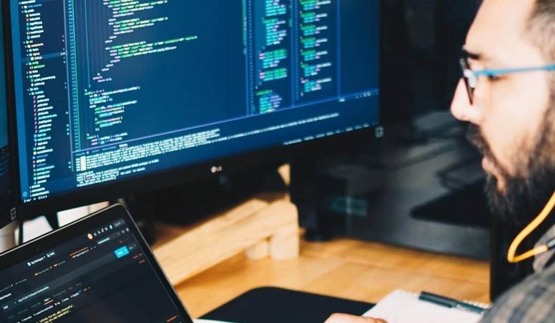 A person works on a laptop with code displayed on the screen, while a large monitor showcases detailed lines of code. The workspace is tech-oriented and focused, seamlessly integrating into the broader IT infrastructure.-ux-design-studio-VzJjPuk53sk-unsplash-scaled-p-1080-1