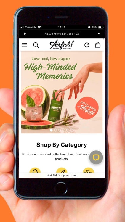 A person holds a smartphone displaying a webpage from Airfield Supply Co., seamlessly integrating their digital transformation strategy. The screen shows a green drink can, a watermelon slice, and the text 