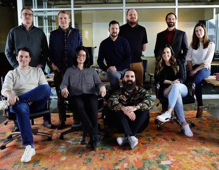 A group of ten individuals, consisting of six men and four women, are posing for a group photo in an office setting typical of software companies in Minneapolis. Five are seated, and the rest stand behind them. They are casually dressed and smiling. The office space includes a colorful rug and visible workspaces.