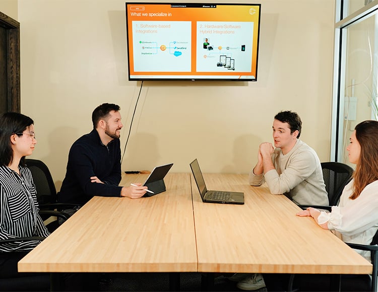 Four people are seated around a conference table, two on each side. Two laptops are open, displaying slides about 