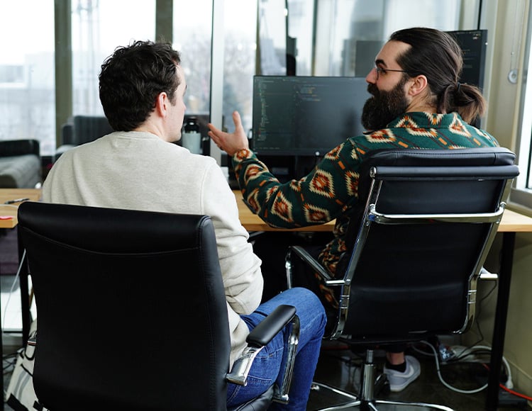 Two men are sitting in office chairs at a desk with computer screens. Engaged in discussion, one gestures while speaking about AI strategy consulting. Their bright room with large windows is filled with focus and innovation.