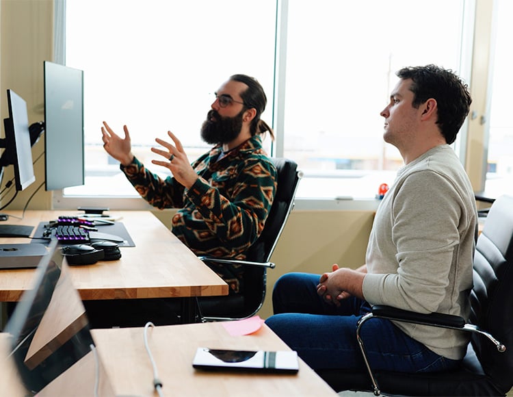 Two people sit at a wooden desk with computers in an office setting, discussing custom software development. One gestures animatedly as they speak, while the other listens attentively from black office chairs. Sunlight pours in through a window, illuminating their collaborative space.