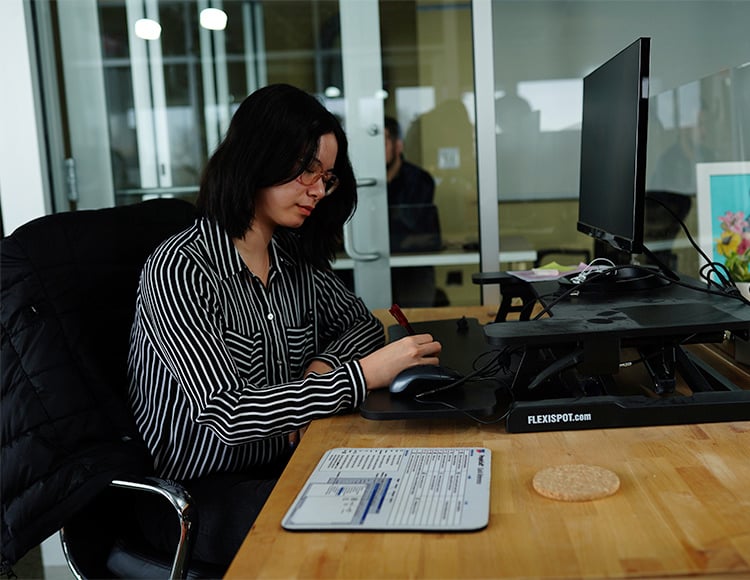 A person with dark hair and glasses is seated at a desk, crafting notes on paper. Dressed in a black and white striped shirt, they face a computer monitor. The office, framed by glass walls, hints at a tech-savvy atmosphere focused on data integration services.