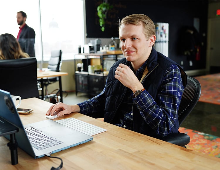 A man in a blue checkered shirt and dark vest sits at a desk, studying custom software development solutions on his laptop. He appears thoughtful, holding his chin. The modern office setting buzzes with colleagues in the background and various office items around.