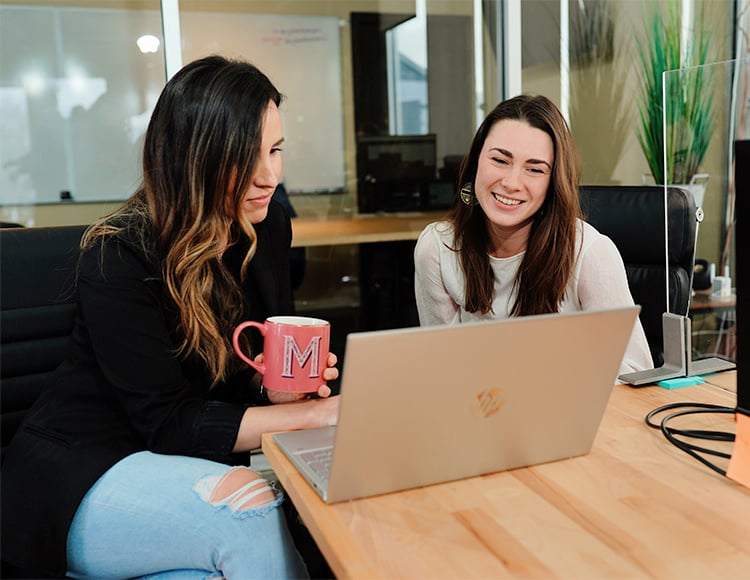 Two women sitting at a wooden desk in an office, smiling and looking at a laptop. One holds a red mug with the letter 
