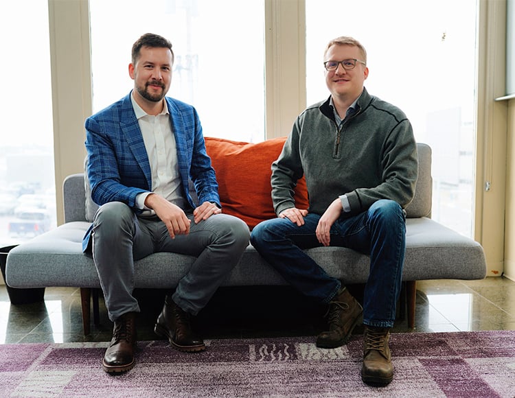Two men, likely colleagues at software companies in Minneapolis, are sitting on a sofa in a well-lit room. The man on the left is sporting a blue checkered blazer and brown shoes, while the man on the right wears a gray sweater and boots. An orange cushion is behind them on the sofa.