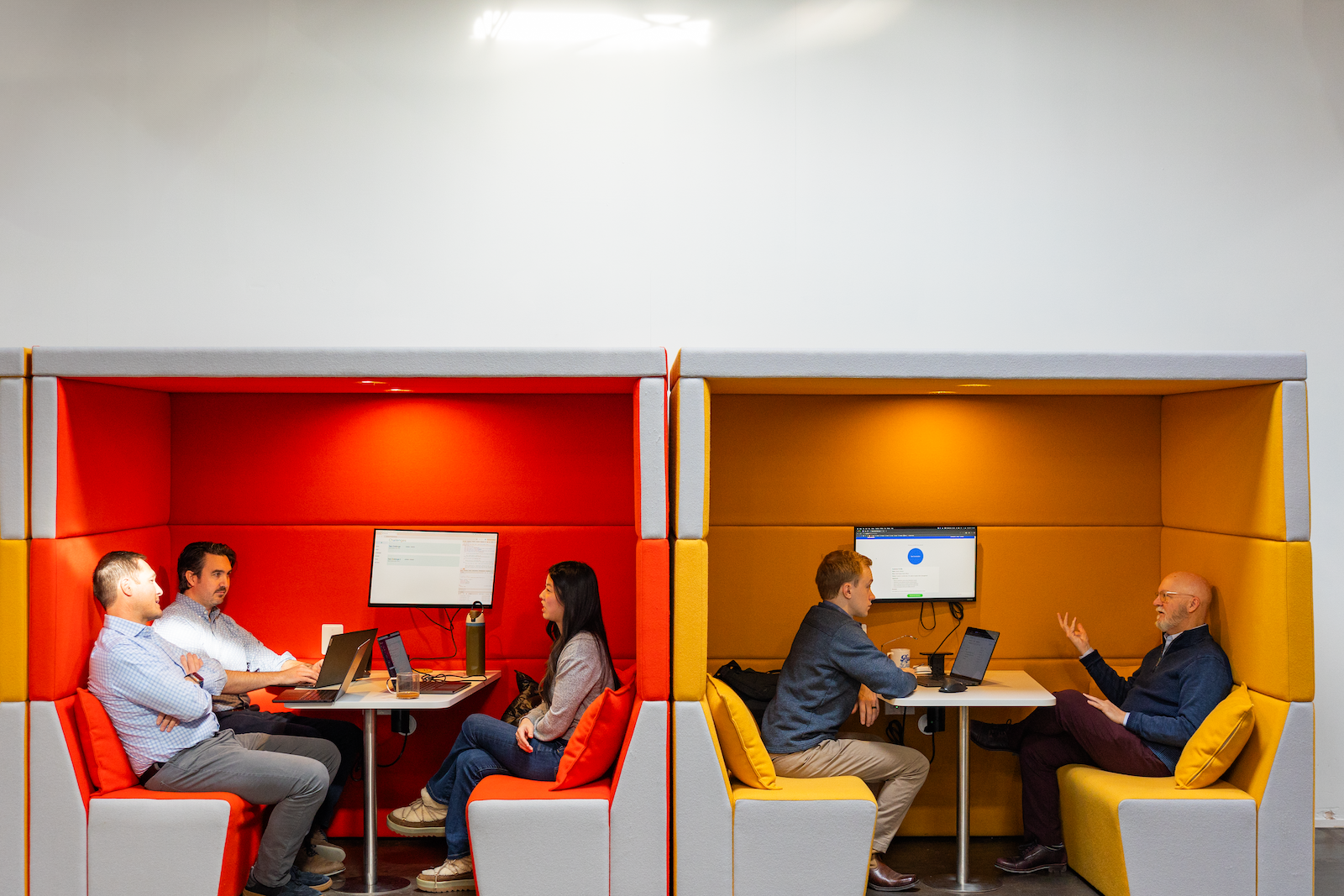 Two groups of people sit in modern, colorful office booths with laptops and monitors, discussing private equity tech solutions. The booths are red and yellow, and each group consists of two men and one woman or two men.