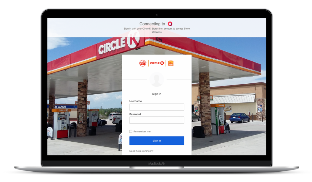 A laptop screen displays a web page for signing into Circle K’s portal, highlighting its robust IT infrastructure with secure username and password fields. In the background, a Circle K gas station is visible, featuring fuel pumps and a car wash area under a red and white canopy.